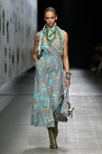 Model walking a runway in a gray floral maxi dress with teal and coral embroidery, paired with a teal scarf and olive boots, carrying a gray handbag.