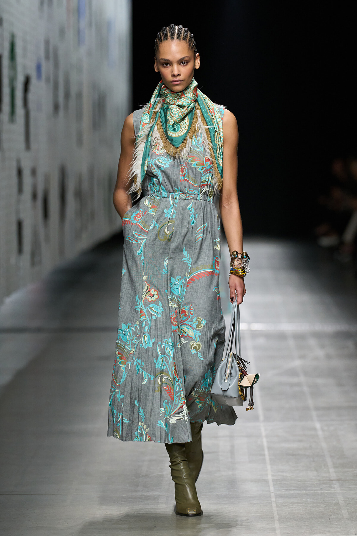 Model walking a runway in a gray floral maxi dress with teal and coral embroidery, paired with a teal scarf and olive boots, carrying a gray handbag.