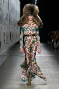 Model on runway in a long patterned dress with green, burgundy, and cream swirls, belt, and an oversized fur hat of matching color.