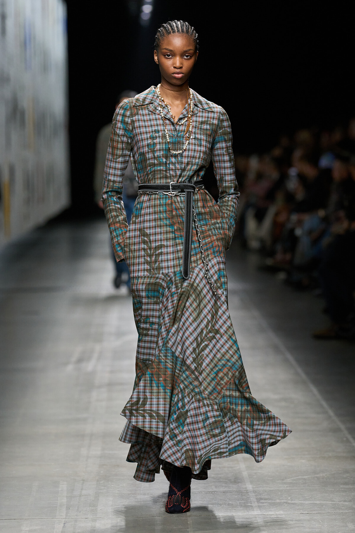 Model walks the runway in a floor-length plaid dress with a black belt and braided chain accessory.