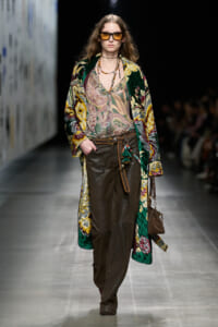 Model struts a fashion runway in a vibrant quilted coat over a paisley blouse and brown leather pants, accessorized with sunglasses and layered necklaces.