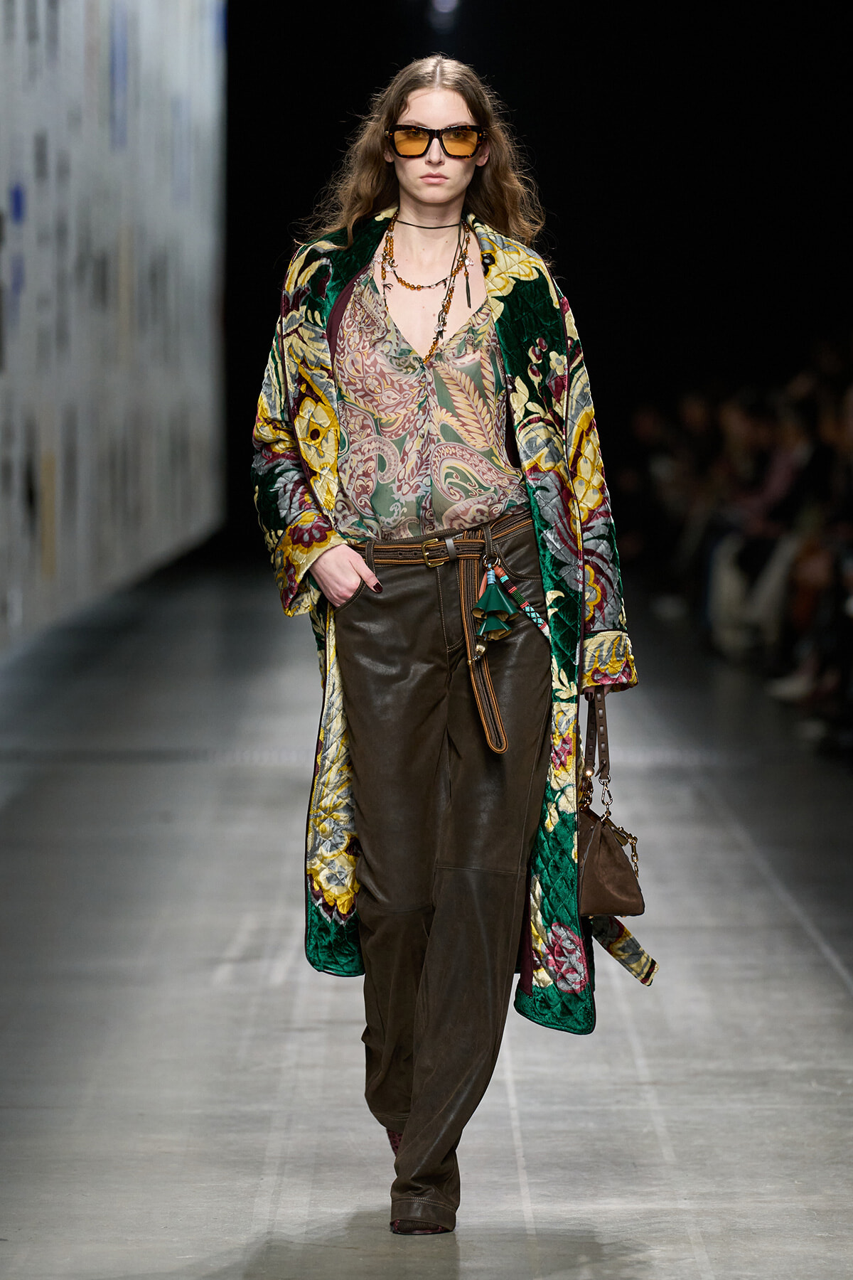 Model struts a fashion runway in a vibrant quilted coat over a paisley blouse and brown leather pants, accessorized with sunglasses and layered necklaces.