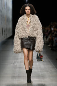 Model on a runway wearing a large beige shaggy faux‑fur coat over a striped blouse and a black leather mini skirt, with dark socks and boots, carrying a small dark handbag with colorful charms.
