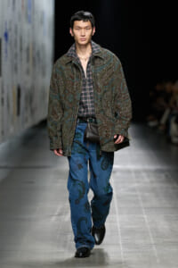 Male model walking runway in a patterned green paisley jacket over a plaid shirt, blue paisley jeans, and black shoes with a small black belt bag.