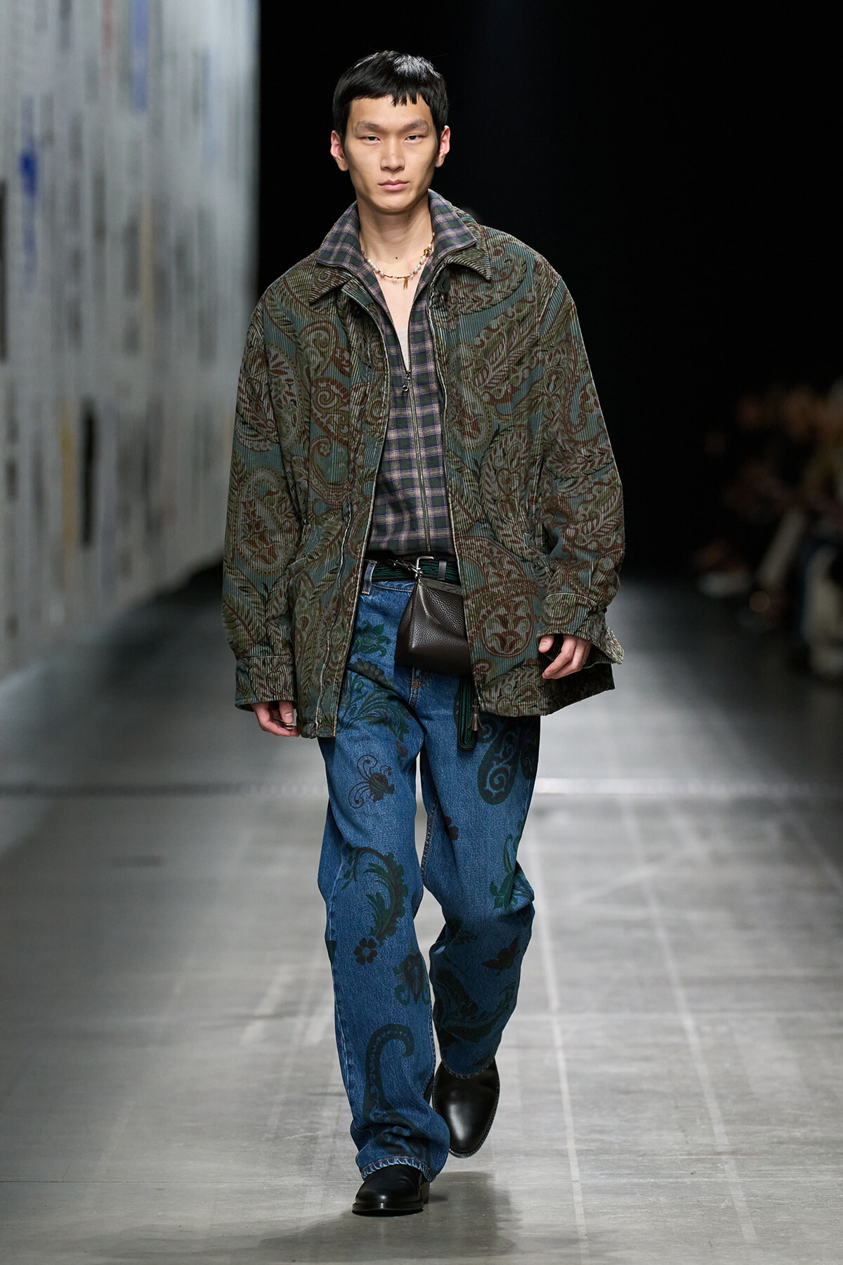 Male model walking runway in a patterned green paisley jacket over a plaid shirt, blue paisley jeans, and black shoes with a small black belt bag.