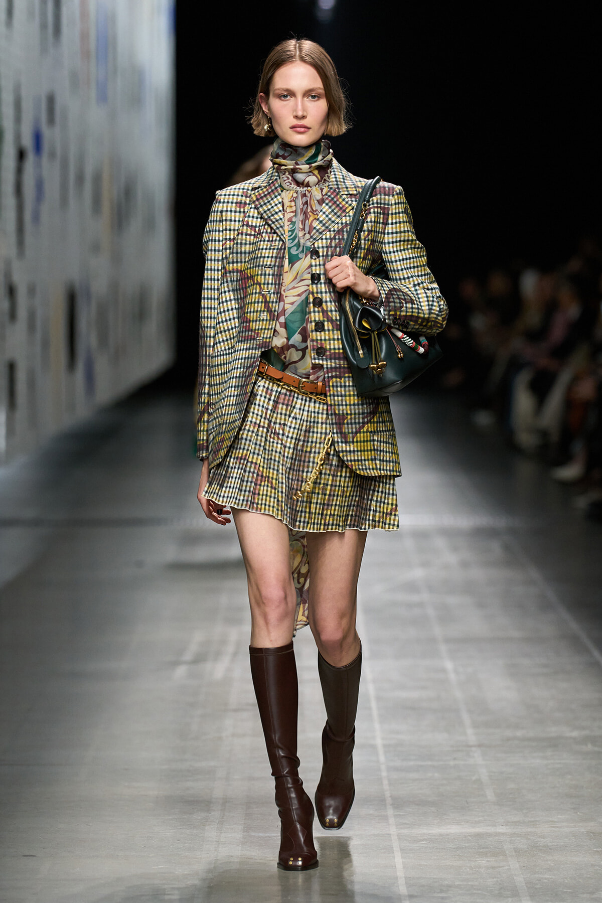 Model strides a runway in a yellow plaid blazer and matching mini skirt, with a patterned scarf and brown knee‑high boots, carrying a dark handbag.