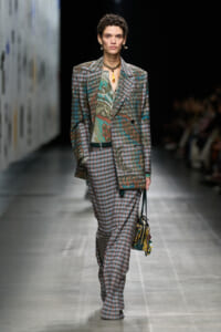 Model on a runway wearing an oversized plaid suit with a paisley-patterned blouse and layered gold jewelry, carrying a patterned handbag.