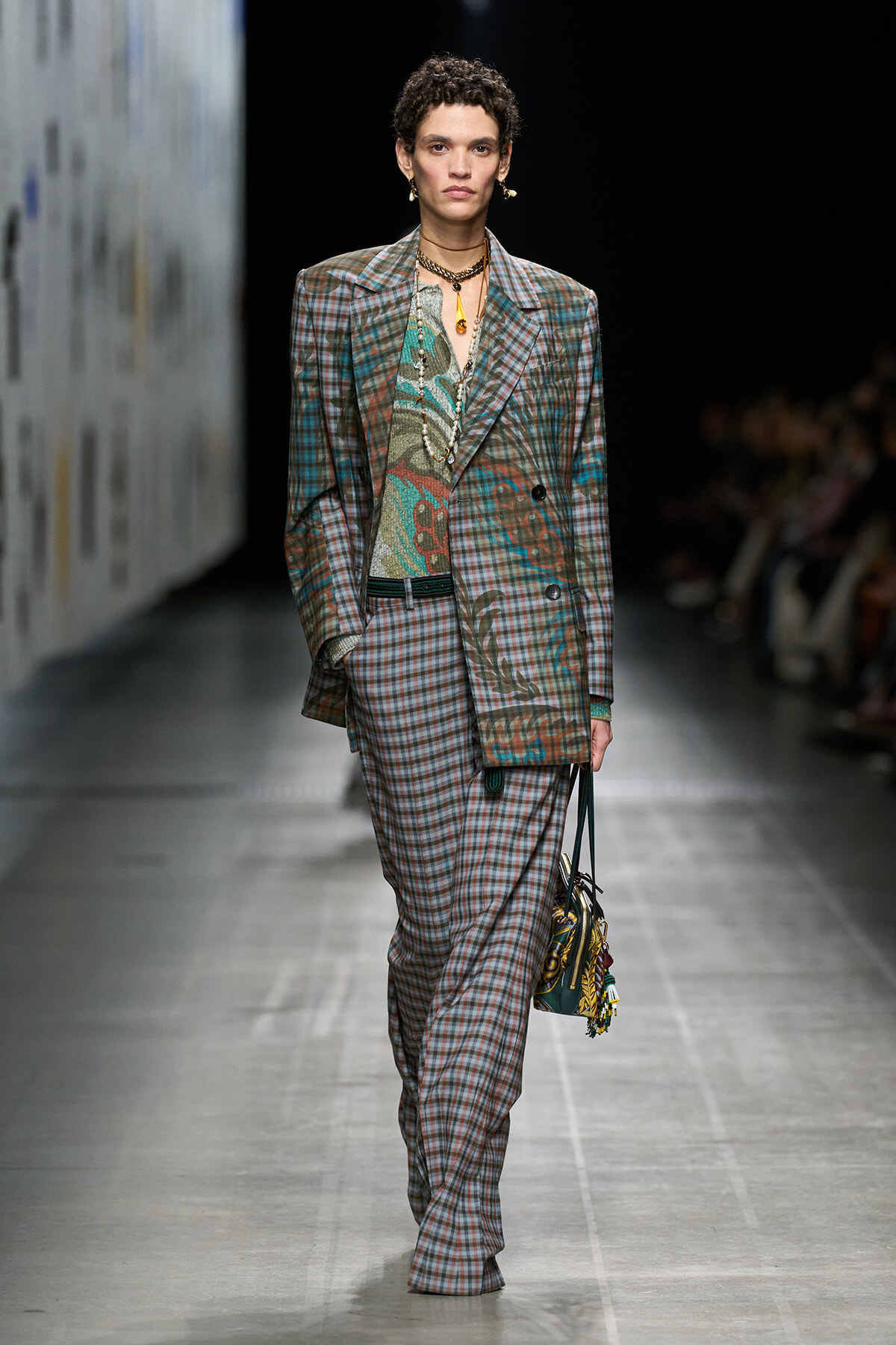 Model on a runway wearing an oversized plaid suit with a paisley-patterned blouse and layered gold jewelry, carrying a patterned handbag.