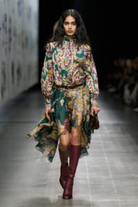 Model walking runway in a colorful floral-printed dress with high neck and long sleeves, cinched with a belt, burgundy knee-high boots, and a small handbag.