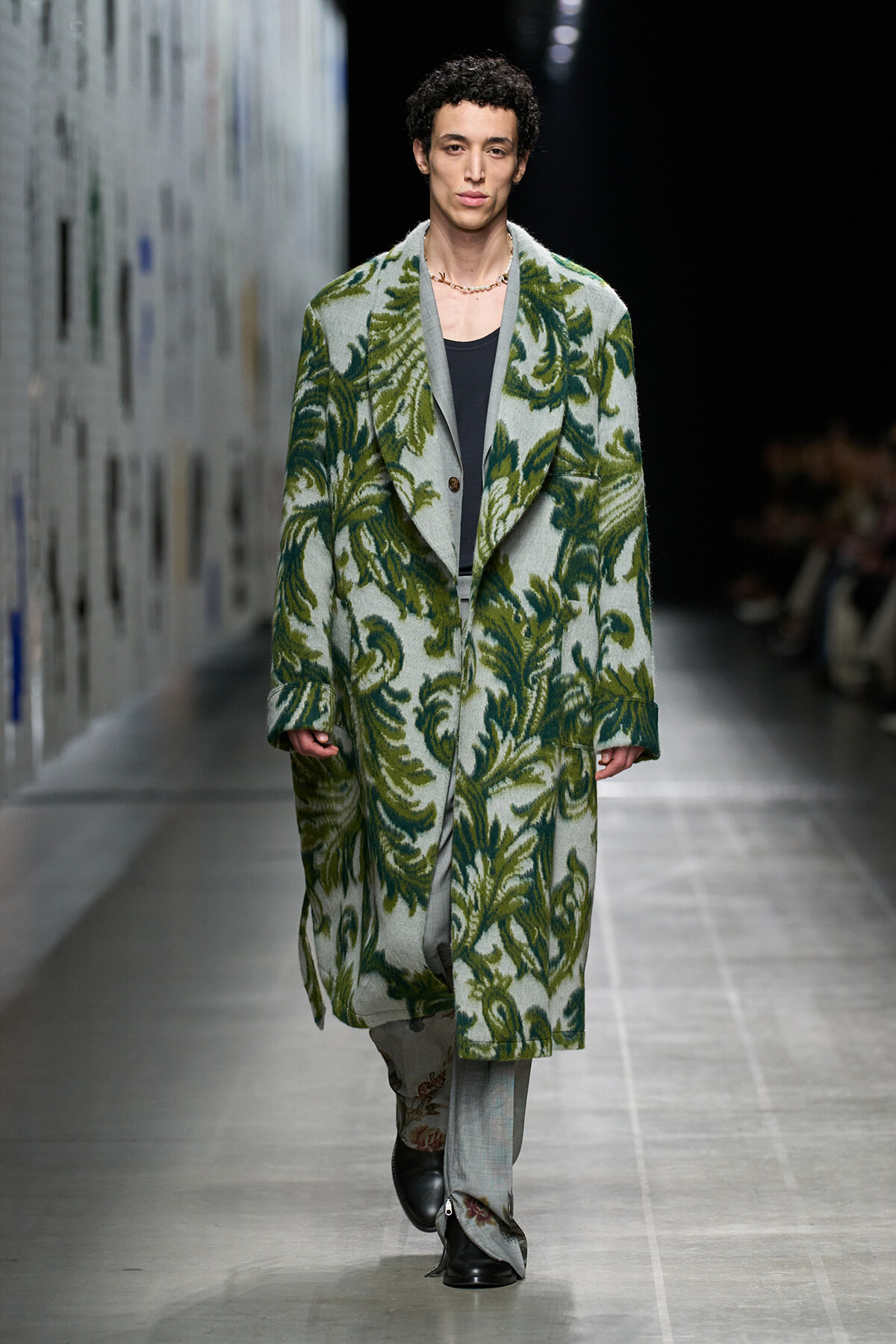 Male model wearing an oversized green fern-patterned coat walks the runway in a dark shirt beneath.