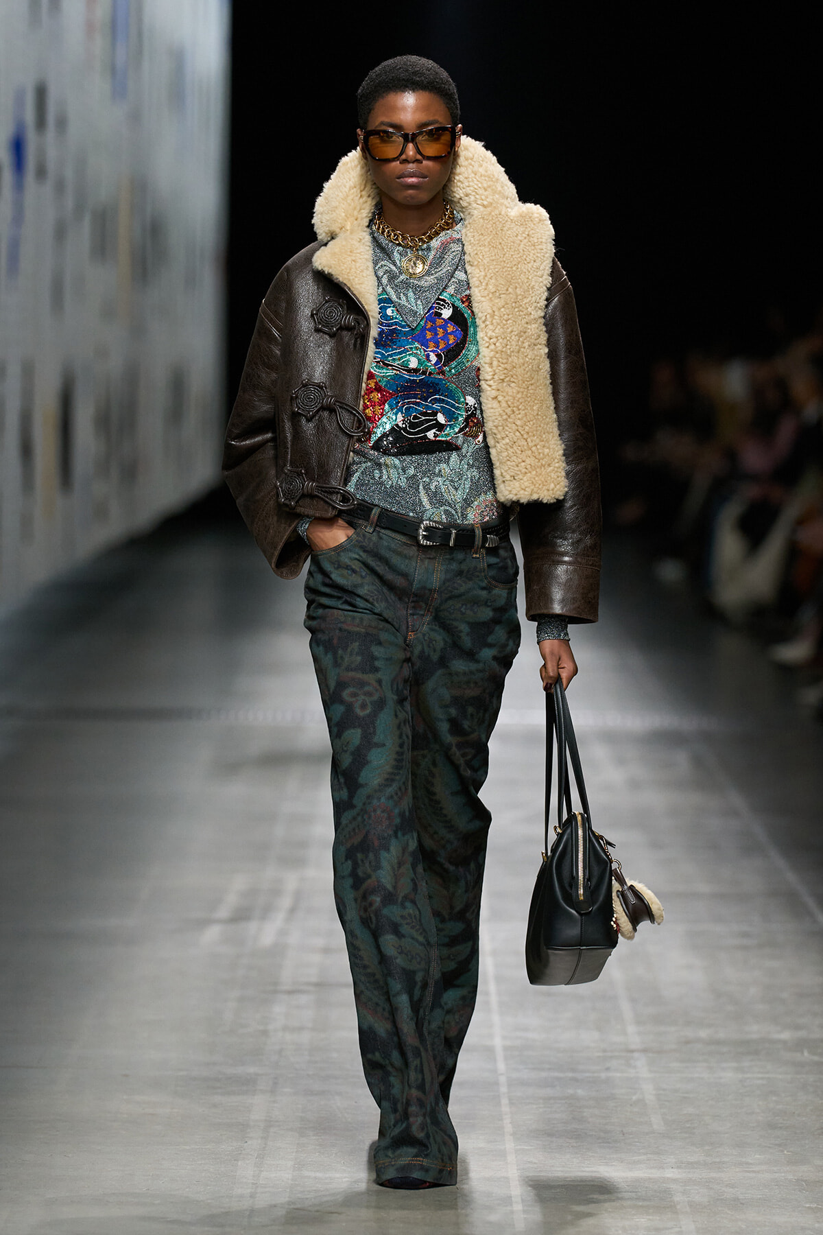 Model walking a runway in a dark brown shearling-lined leather jacket over a colorful patterned top, paired with green paisley pants and chunky gold jewelry, wearing brown sunglasses and carrying a black handbag with a fur keychain.