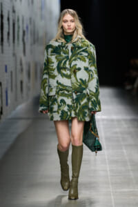 Model walking a runway in a green, palm- and fern-patterned coat with green turtleneck, olive knee-high boots, and a green handbag.