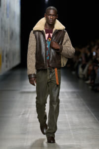 Male model walking runway in a brown shearling-lined leather jacket, olive cargo pants, and dark boots.”