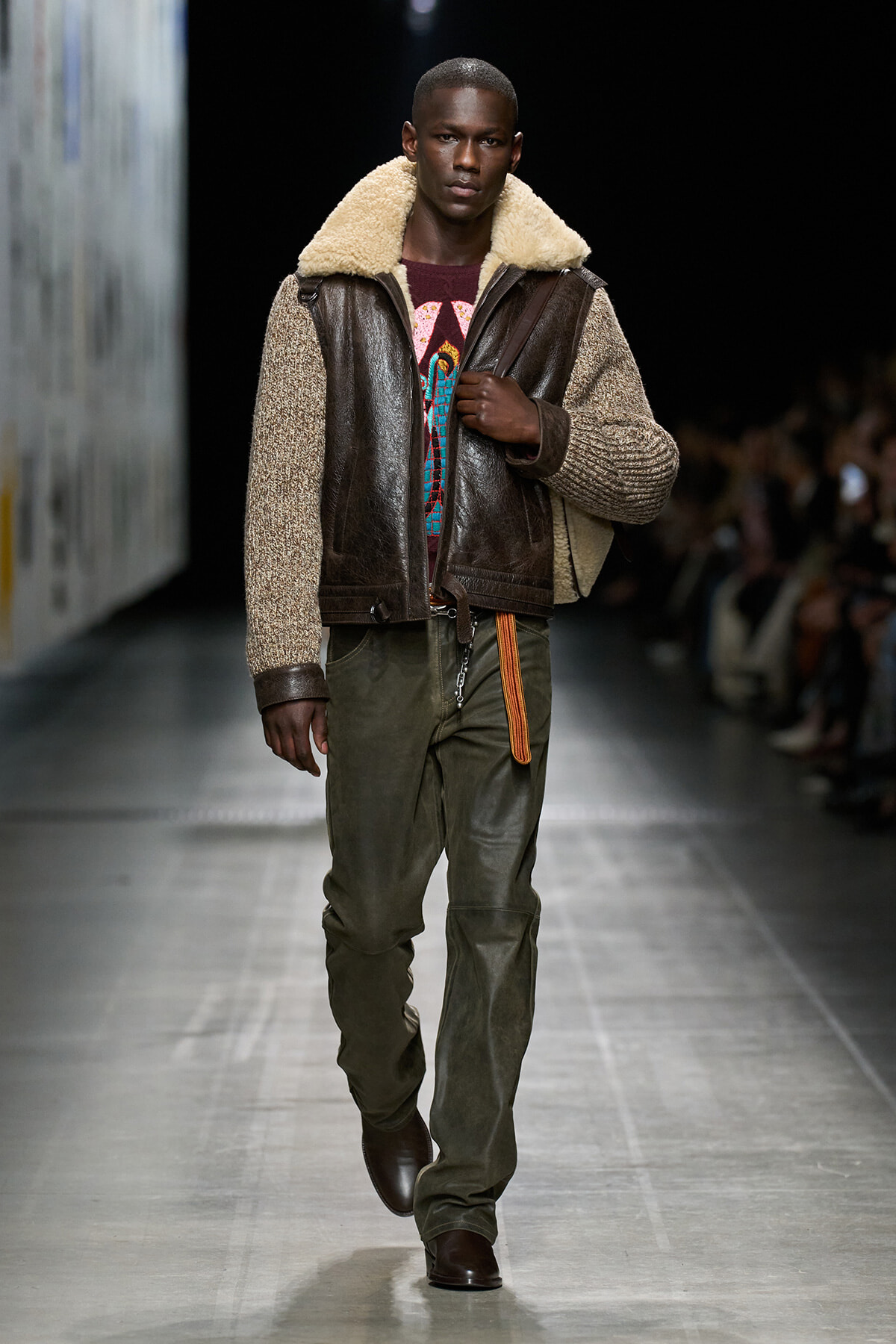 Male model walking runway in a brown shearling-lined leather jacket, olive cargo pants, and dark boots.”