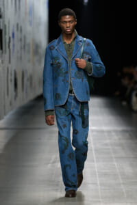 Male model walks the runway in a blue denim suit with green floral embroidery, carrying a green shoulder bag.