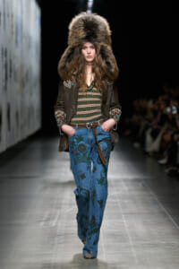Model walks the runway wearing a large fur hat, embroidered brown jacket, striped top, and floral blue jeans with a belt.