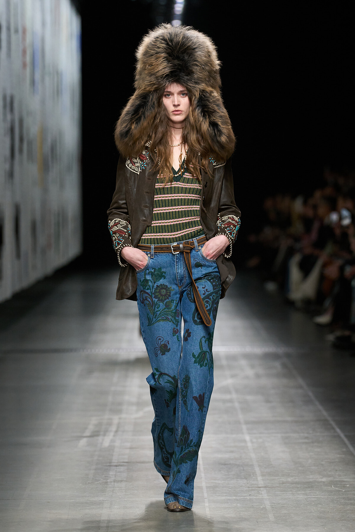 Model walks the runway wearing a large fur hat, embroidered brown jacket, striped top, and floral blue jeans with a belt.