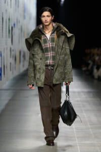Model wearing a green quilted coat with fur collar over plaid shirt and brown trousers, carrying a black handbag on a runway.
