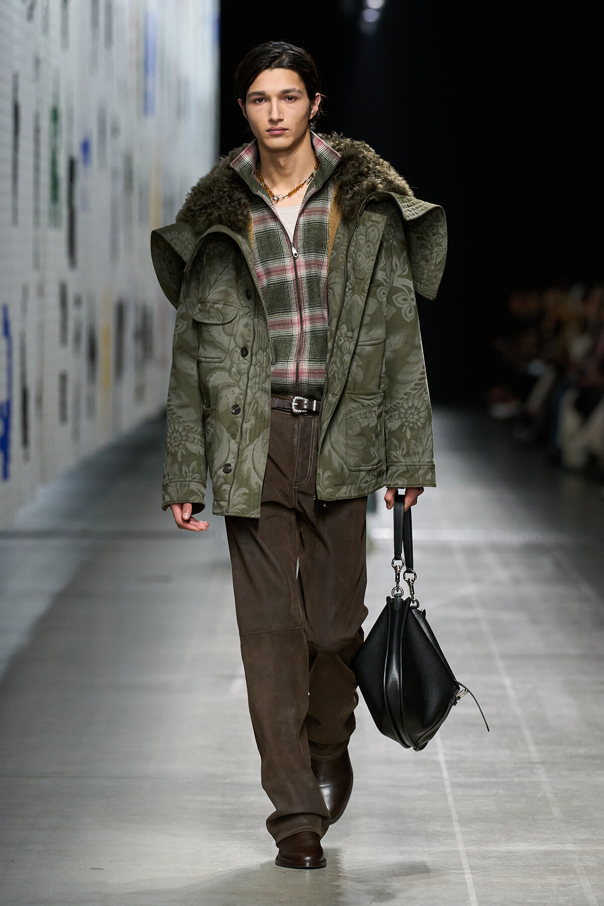Model wearing a green quilted coat with fur collar over plaid shirt and brown trousers, carrying a black handbag on a runway.