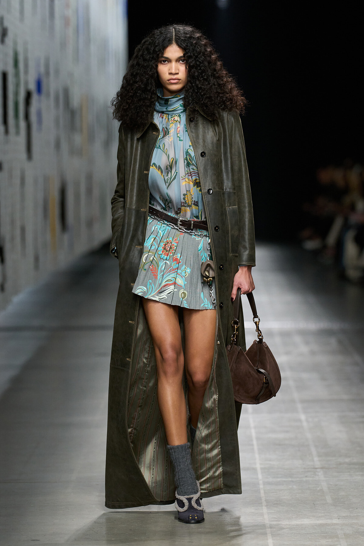 Model walking a runway in a long olive-green coat over a pastel floral mini dress, cinched with a belt, carrying a brown suede handbag.