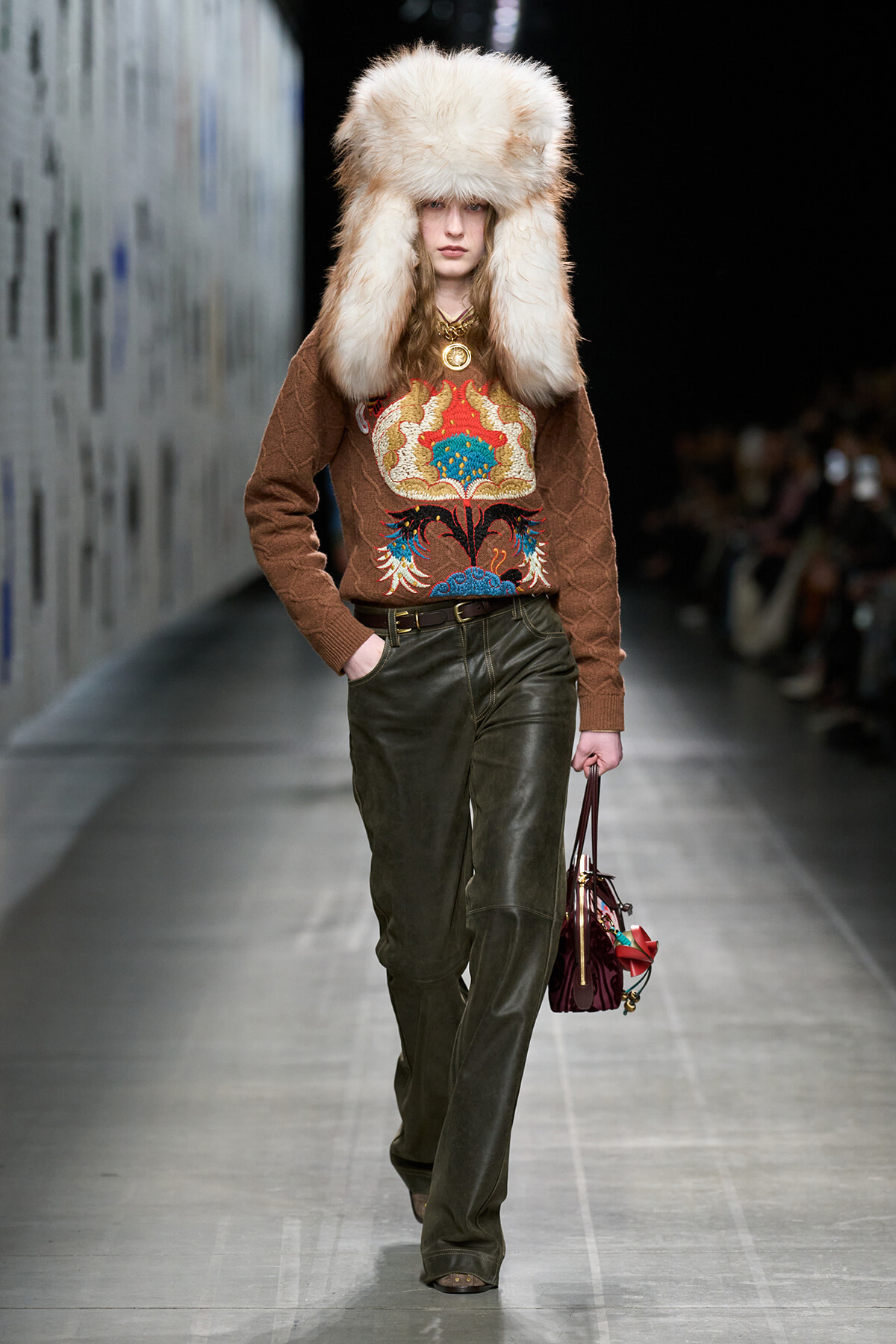 Model walks the runway in a brown patterned sweater with bold embroidery, olive leather pants, and a large fluffy fur hat, carrying a burgundy handbag.