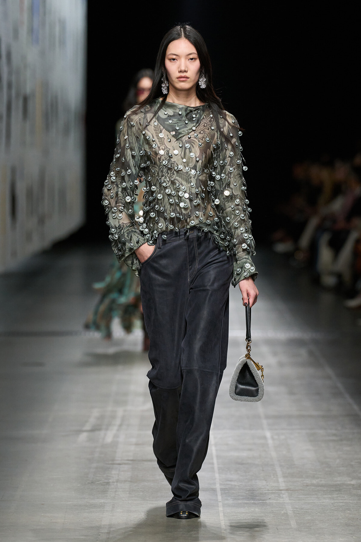 Model walking runway in sheer, sequined gray-green blouse with dangling discs, paired with loose dark trousers and a small gray handbag.