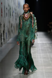 Model on a runway wearing a sheer emerald green, paisley-printed gown with a decorative scarf draped around the neck and brown ankle boots.