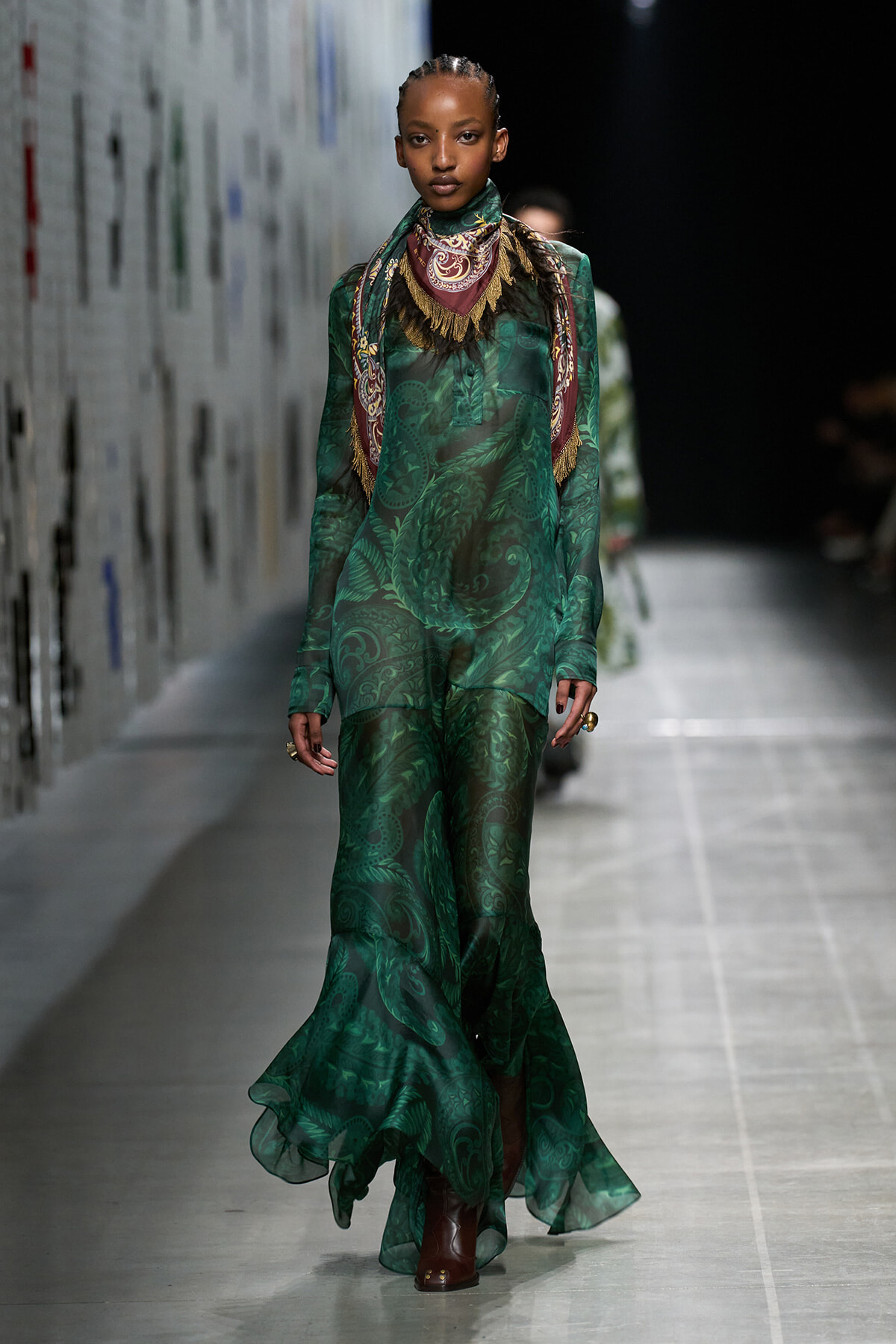 Model on a runway wearing a sheer emerald green, paisley-printed gown with a decorative scarf draped around the neck and brown ankle boots.