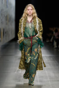 Model walks a fashion runway in a green floral print jumpsuit with a long, textured yellow-brown coat and layered necklaces.
