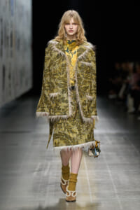 Model walks a fashion runway wearing a textured yellow‑green coat with white fur trim over a patterned yellow dress and mustard socks with wooden sandals, holding a small bag.