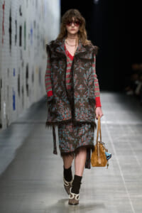 Model walking a runway in a textured plaid coat with fur trim over a red top and matching skirt, sunglasses, and a tan handbag.