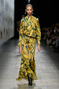Model walks the runway in a long green-and-yellow floral print dress with a black belt and statement earrings.