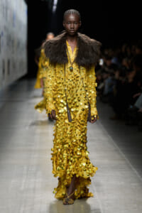 Model on a fashion runway wearing a yellow gown densely covered in gold coin-like sequins, with a dark fur stole and long pendant necklace.