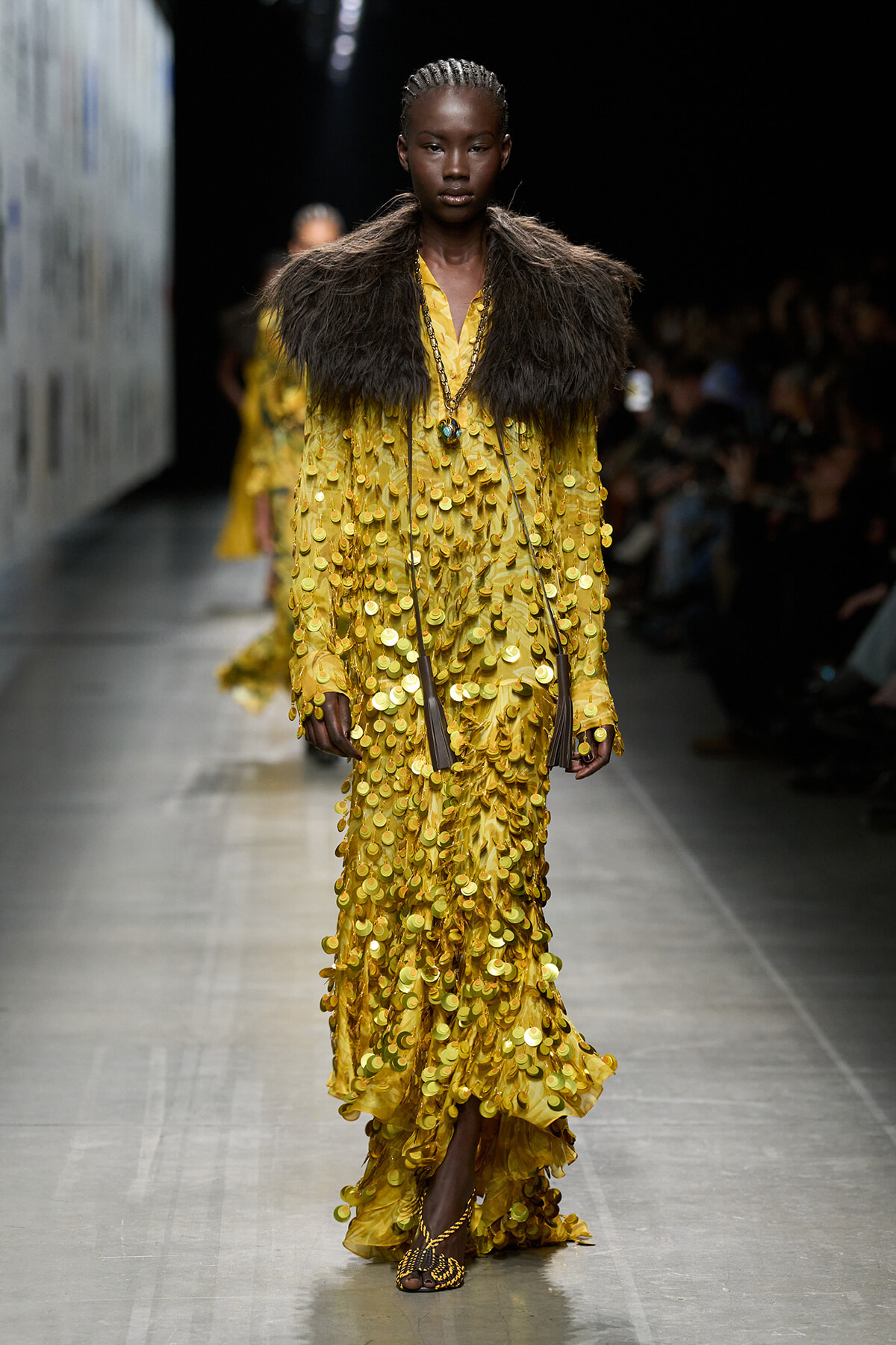 Model on a fashion runway wearing a yellow gown densely covered in gold coin-like sequins, with a dark fur stole and long pendant necklace.
