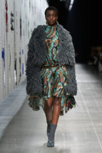 Model on a runway wearing a bold teal and brown floral dress under a large gray shaggy fur coat, accessorized with a belt and earrings.