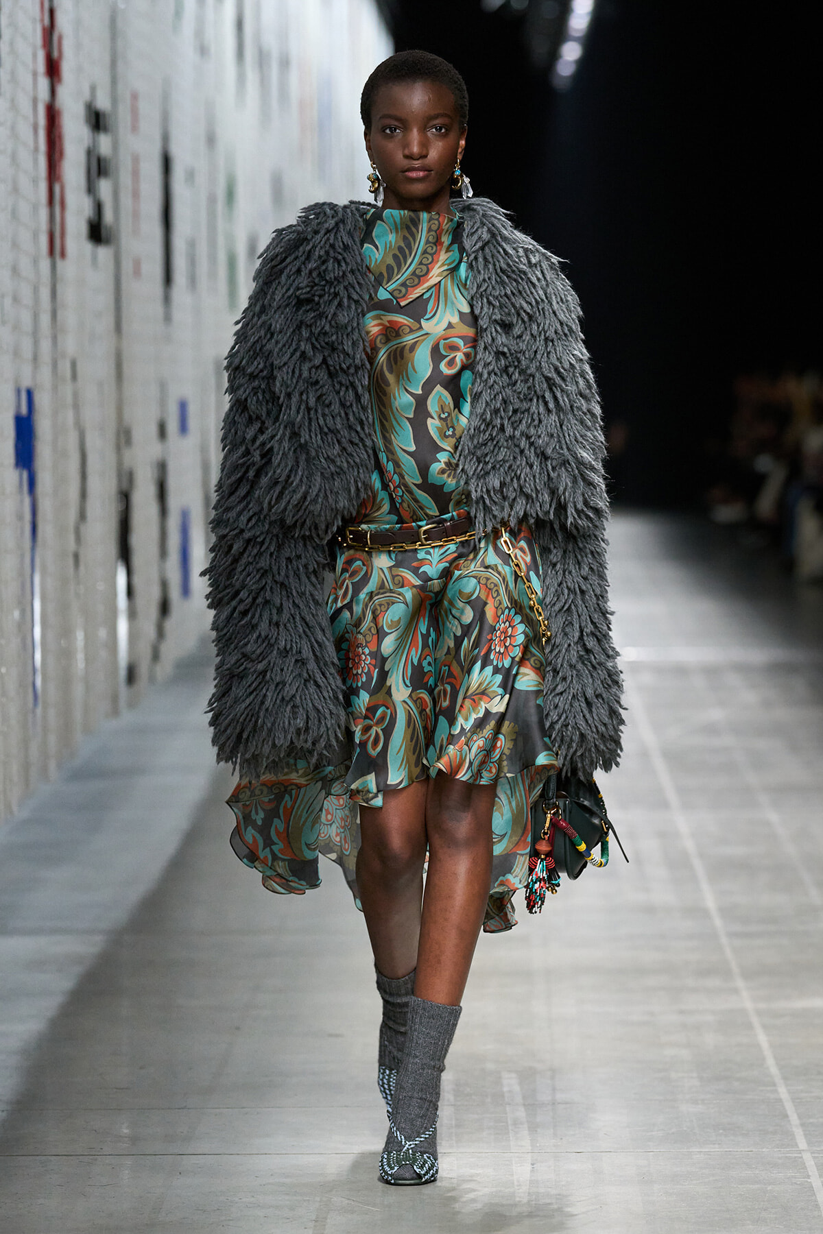 Model on a runway wearing a bold teal and brown floral dress under a large gray shaggy fur coat, accessorized with a belt and earrings.