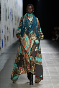 Model walking a runway in a long flowing paisley-patterned dress with teal, brown, and red hues and a blue fringe scarf.