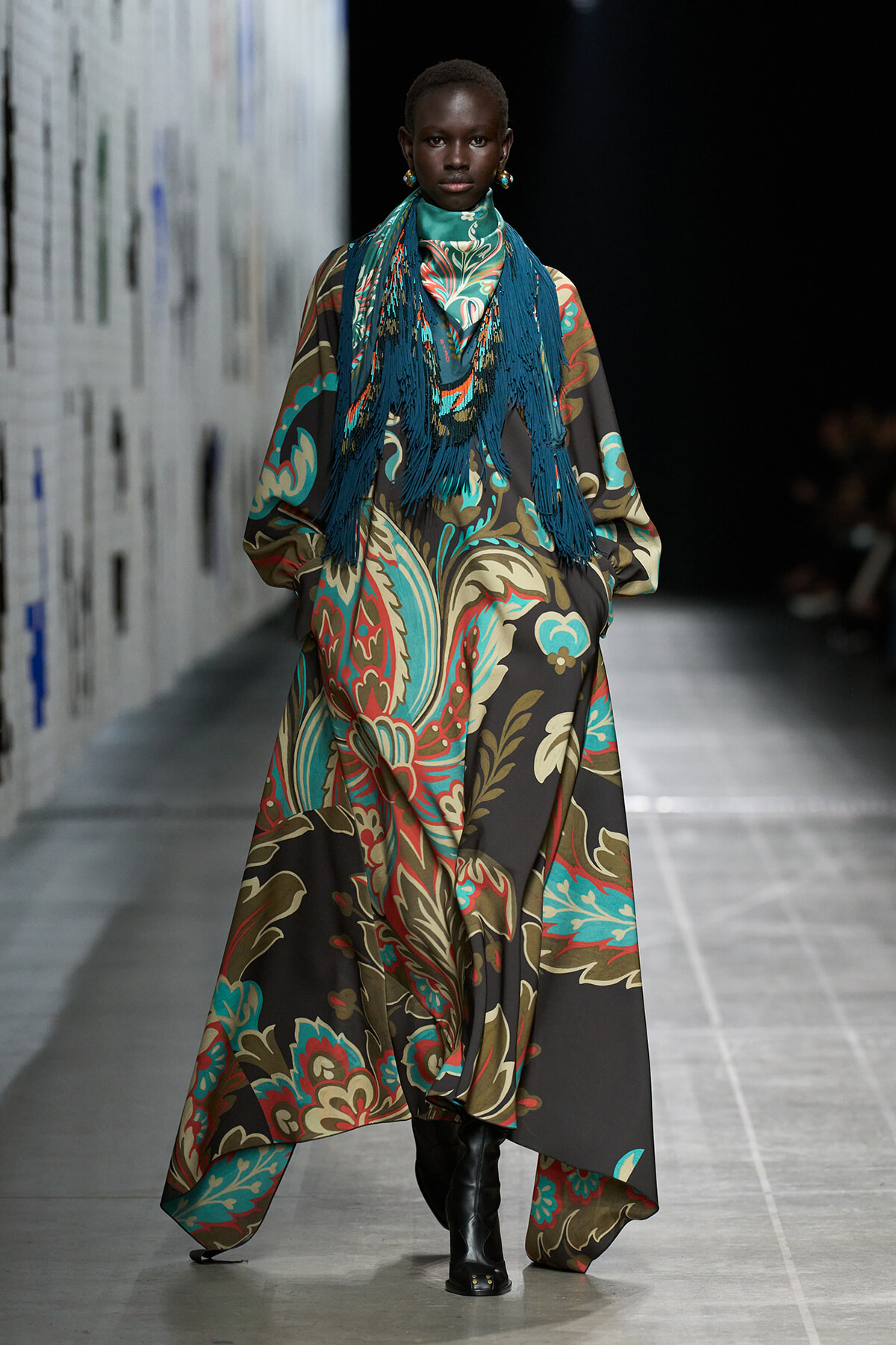 Model walking a runway in a long flowing paisley-patterned dress with teal, brown, and red hues and a blue fringe scarf.