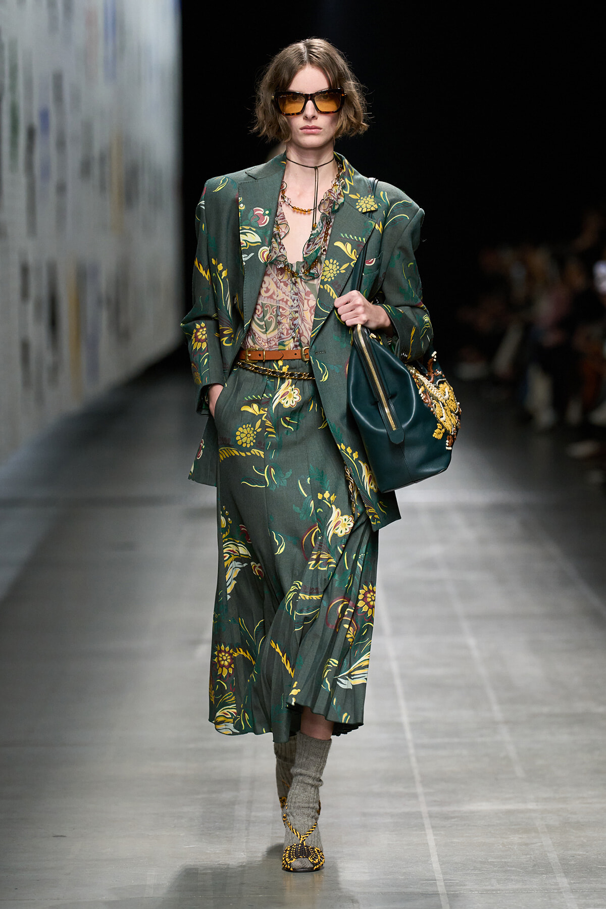 Model on a runway wearing a dark green floral suit with an oversized blazer and matching skirt, brown belt, printed blouse, and large sunglasses, carrying a dark green bag.