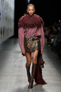 Model walking runway in burgundy fringe cape over a pink striped blouse and a bold floral mini skirt, paired with tall brown boots and a brown handbag.
