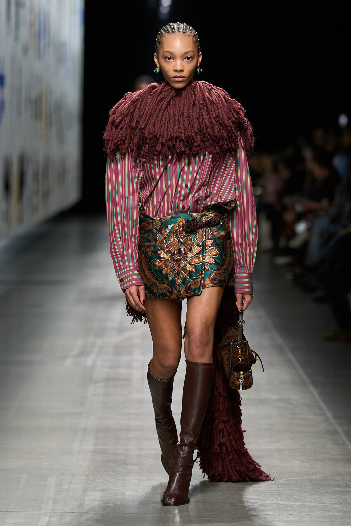 Model walking runway in burgundy fringe cape over a pink striped blouse and a bold floral mini skirt, paired with tall brown boots and a brown handbag.