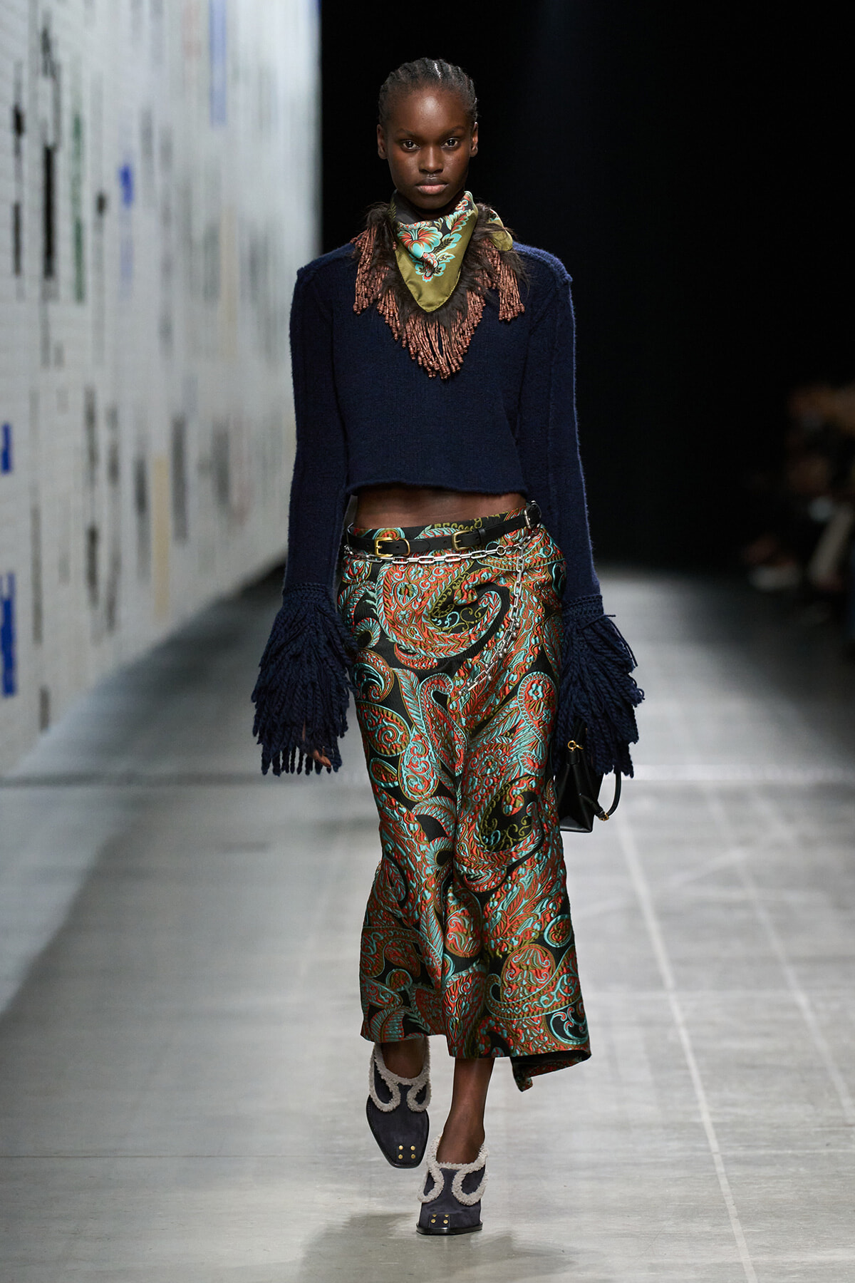Model walking a runway in a navy cropped knit sweater with fringed sleeves, paisley print wide-leg pants, a green scarf, and a belt.