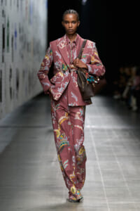 Model walks a runway in a dusty rose floral suit with a matching striped shirt and brown handbag.