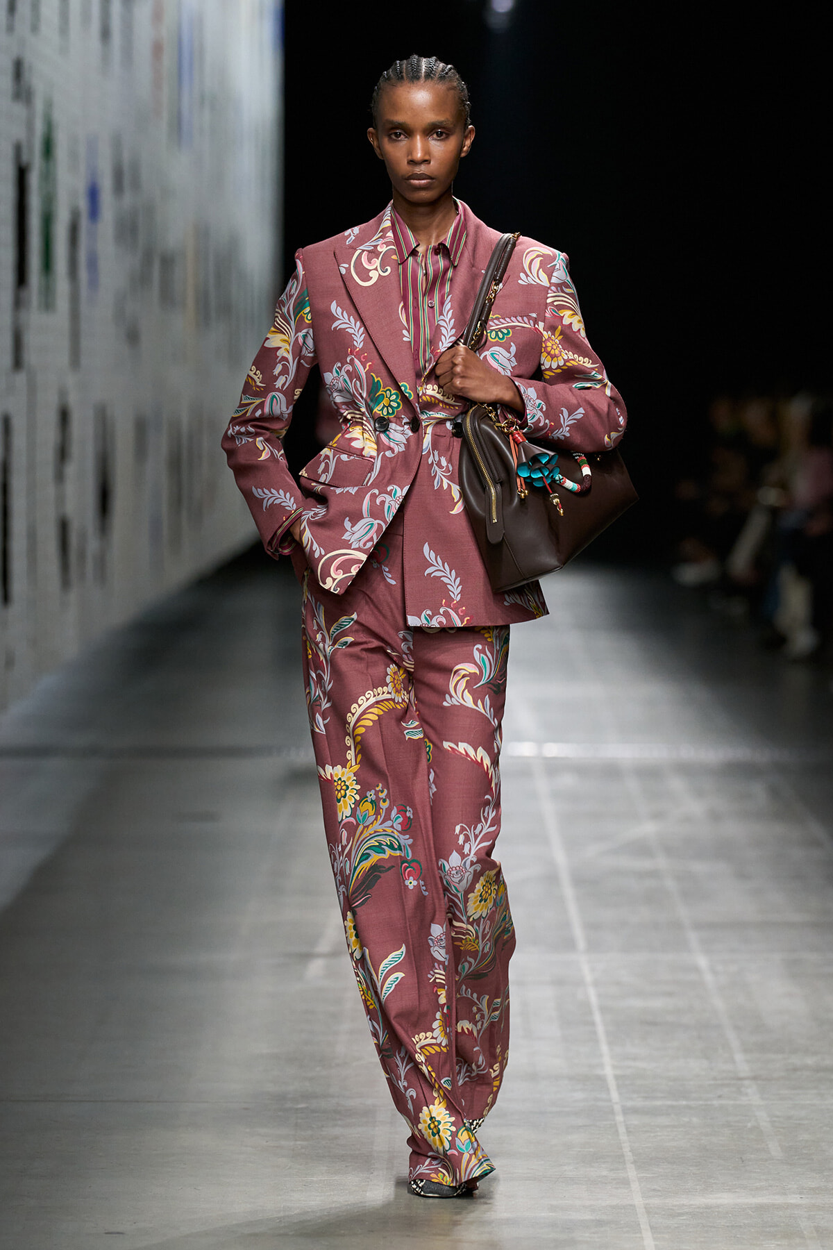 Model walks a runway in a dusty rose floral suit with a matching striped shirt and brown handbag.