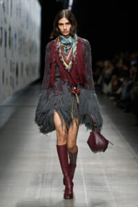 Model on runway wearing a fringed maroon and gray knitted dress with a colorful scarf and burgundy knee-high boots, carrying a burgundy handbag.