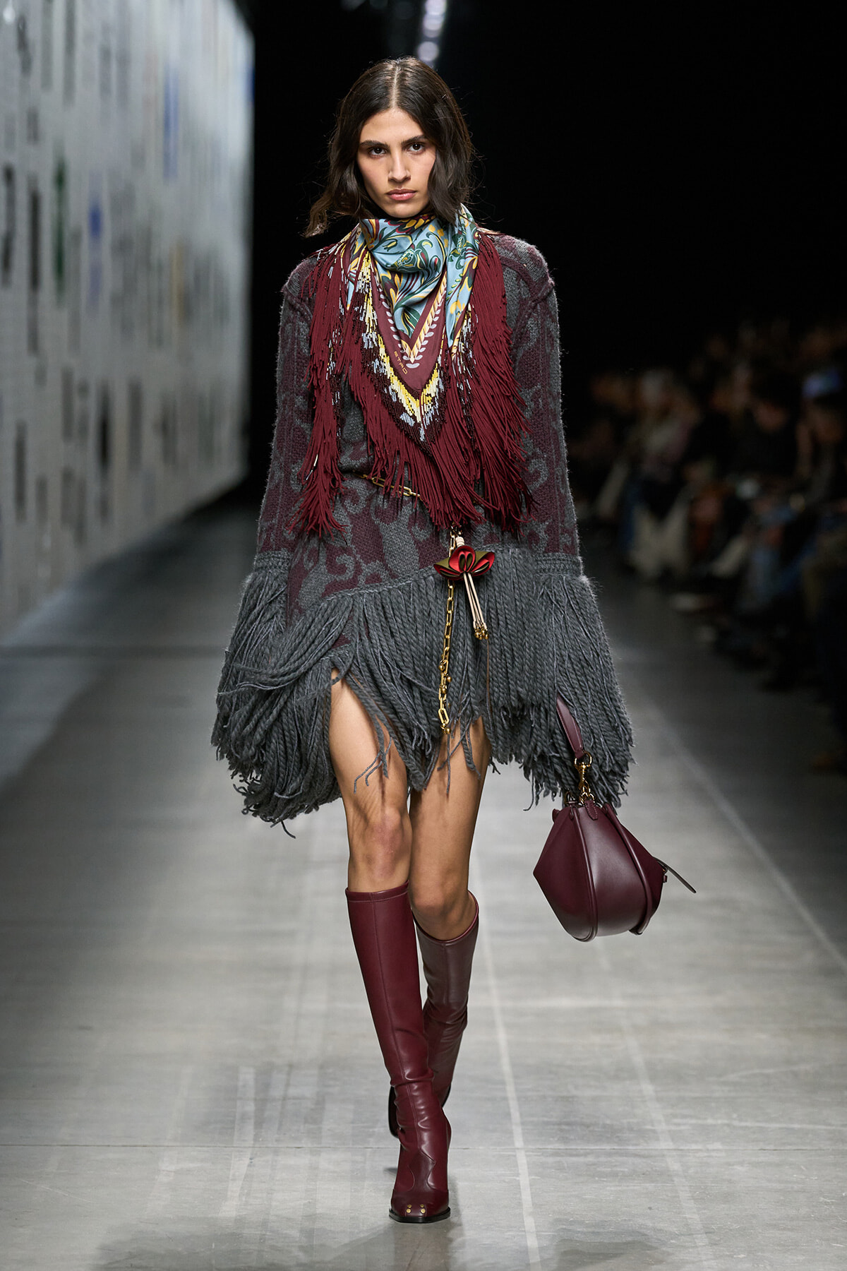 Model on runway wearing a fringed maroon and gray knitted dress with a colorful scarf and burgundy knee-high boots, carrying a burgundy handbag.