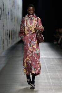 Model walks the runway in a long mauve floral maxi dress with a layered fringe bib, carrying a burgundy shoulder bag.