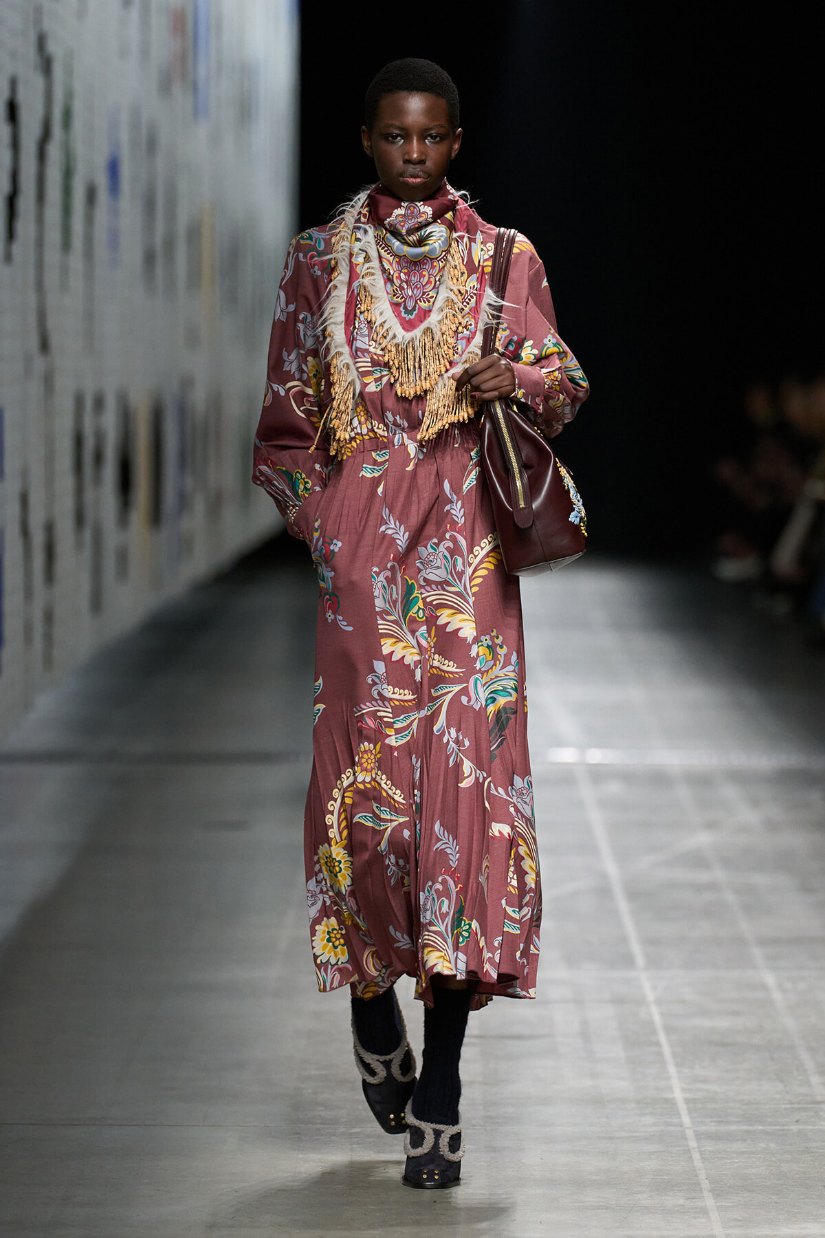 Model walks the runway in a long mauve floral maxi dress with a layered fringe bib, carrying a burgundy shoulder bag.