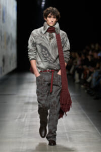 Male model on runway wearing a gray patterned knit sweater, burgundy scarf, gray floral trousers with a brown belt and dark boots.