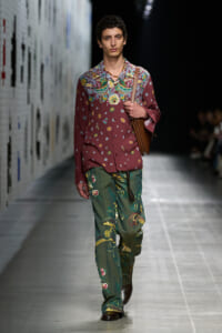 Male model walks a runway in a burgundy floral shirt and green botanical-print pants, carrying a brown crossbody bag.